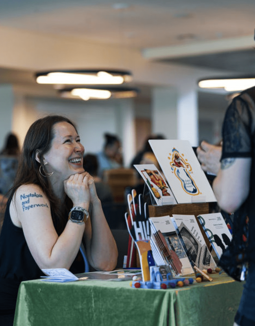 Smiling woman at table with green tablecloth, on top of which are zines and art prints.